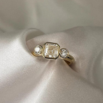 Asscher Cut Trilogy Diamond Engagement Ring featuring a 1.5ct IGI Certified Asscher Cut Lab Grown Diamond with round lab grown diamond side stones in solid gold. Three-stone engagement ring with 1.82ct total carat weight.