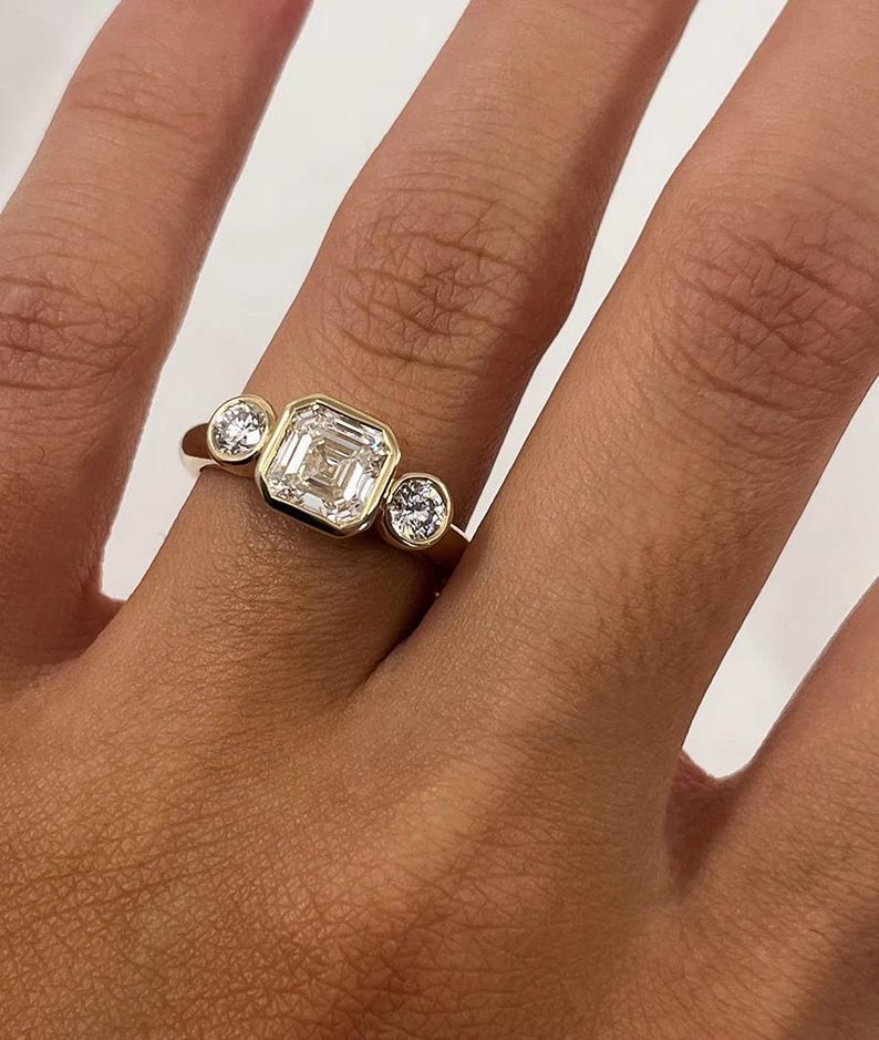 Asscher Cut Trilogy Diamond Engagement Ring featuring a 1.5ct IGI Certified Asscher Cut Lab Grown Diamond with round lab grown diamond side stones in solid gold. Three-stone engagement ring with 1.82ct total carat weight.