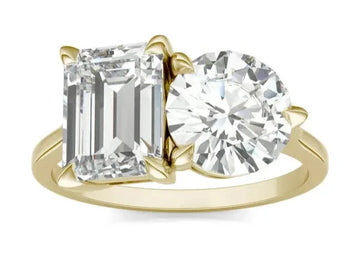 A round brilliant and emerald Toi et Moi engagement ring with a gold band, featuring two emerald stones of different sizes.